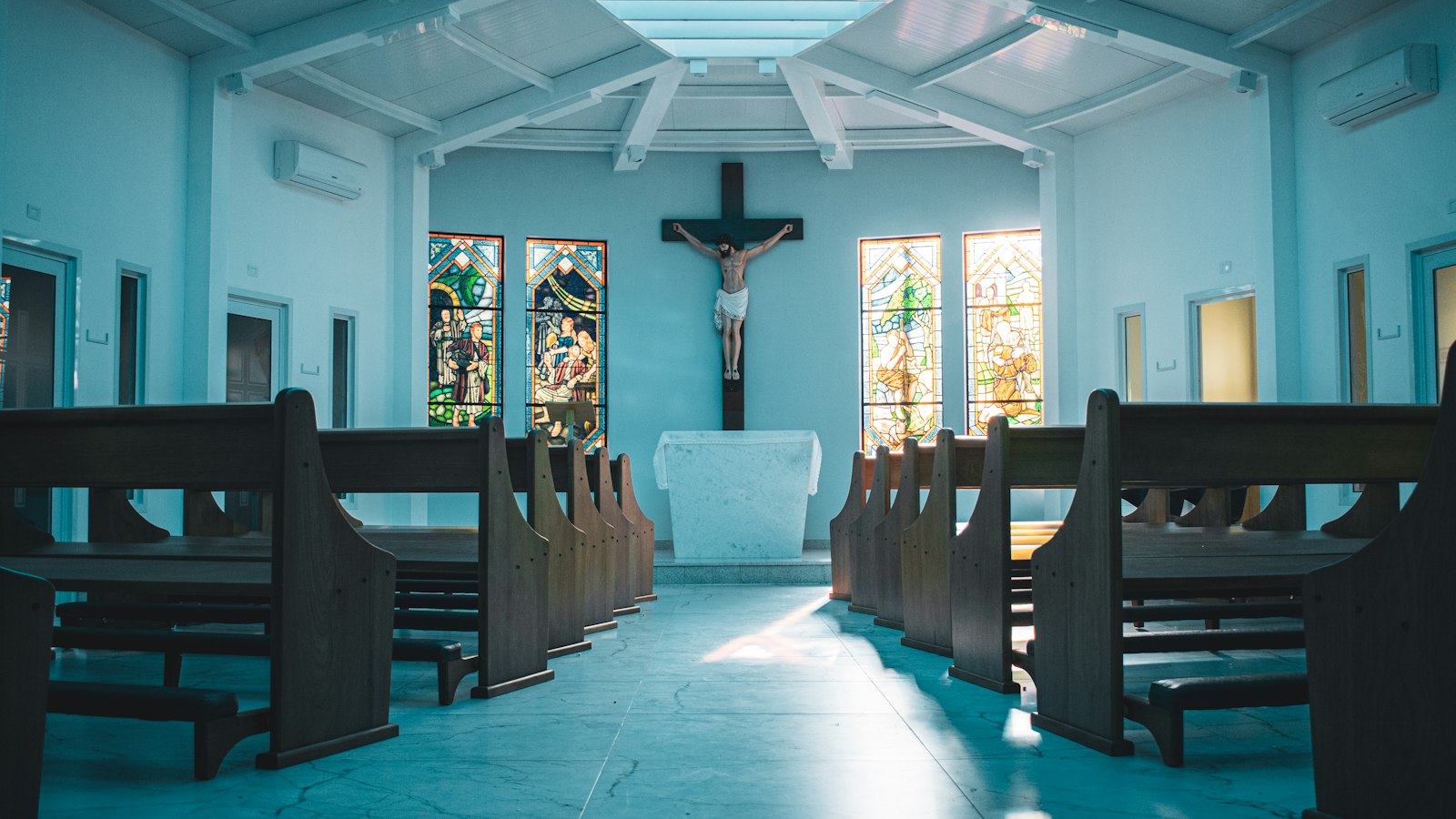 Träkyrkbänkar i ett kyrkorum, ljus genom fönster längs mitgången.