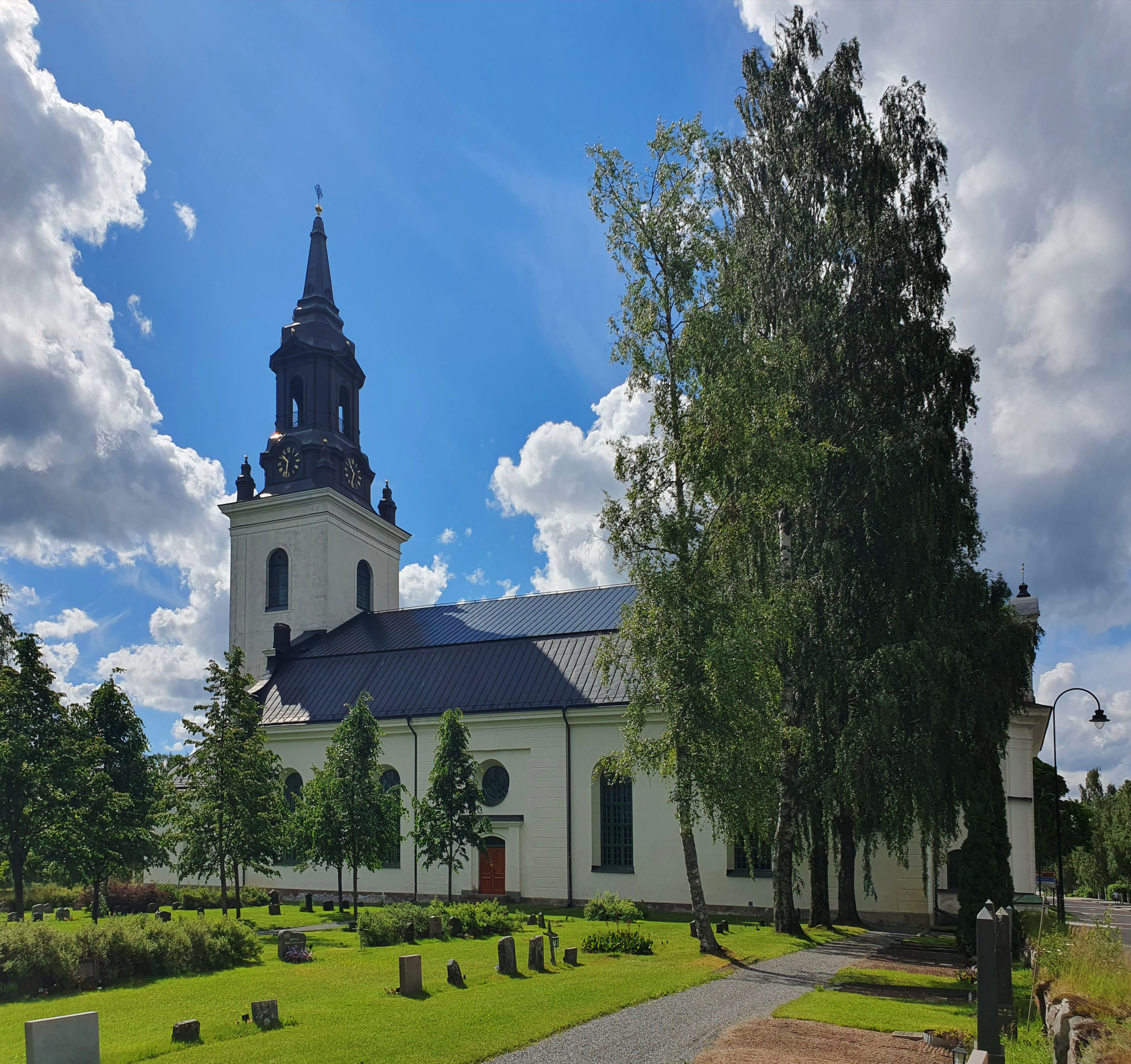 Ockelbo kyrka, ljus kyrka med svart tak, torn och kyrkogård.